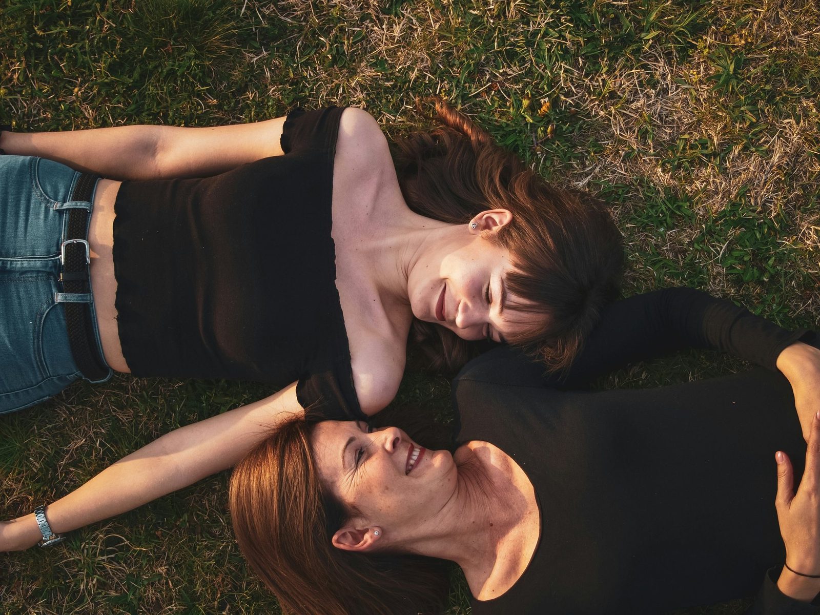 A mother and her adult daughter lying head to head in the grass, smiling at each other in warm evening light.